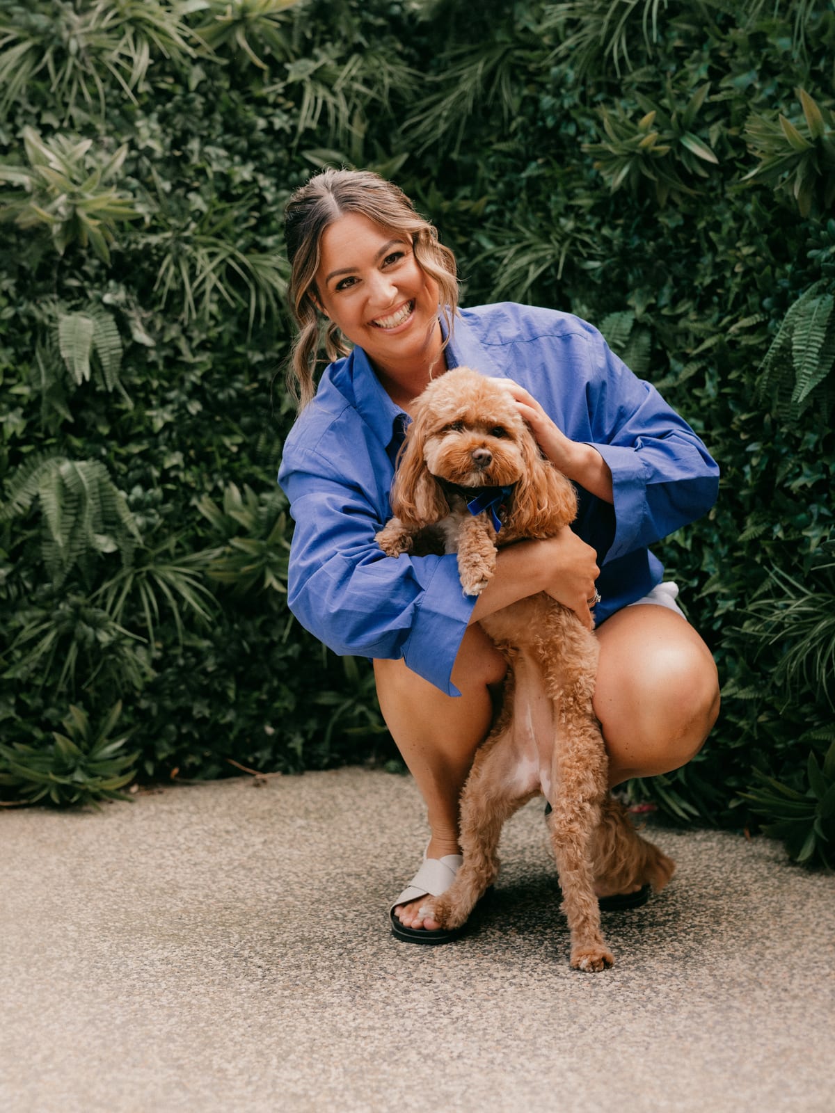 Leah Ferguson crouching in front of a green hedge, smiling with her arms around her cavoodle