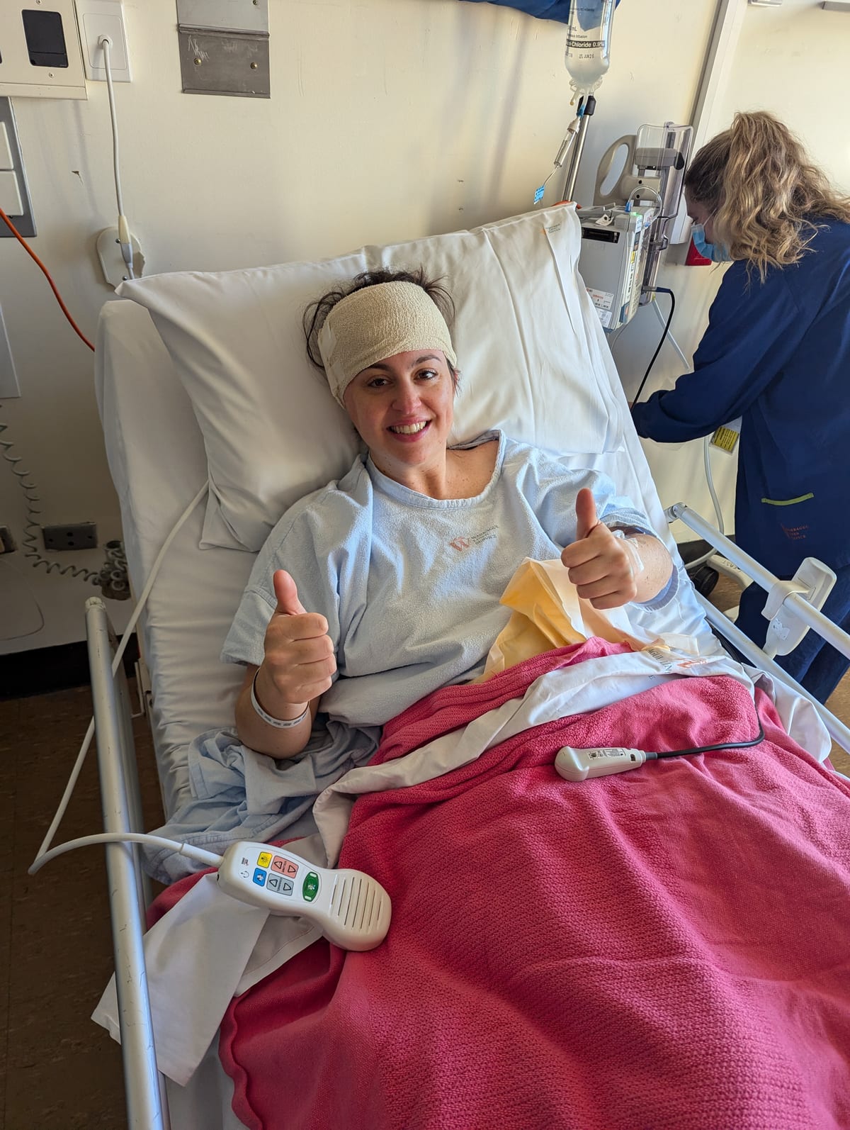 Leah Ferguson in a hospital bed after brain surgery, head bandaged, giving two thumbs up